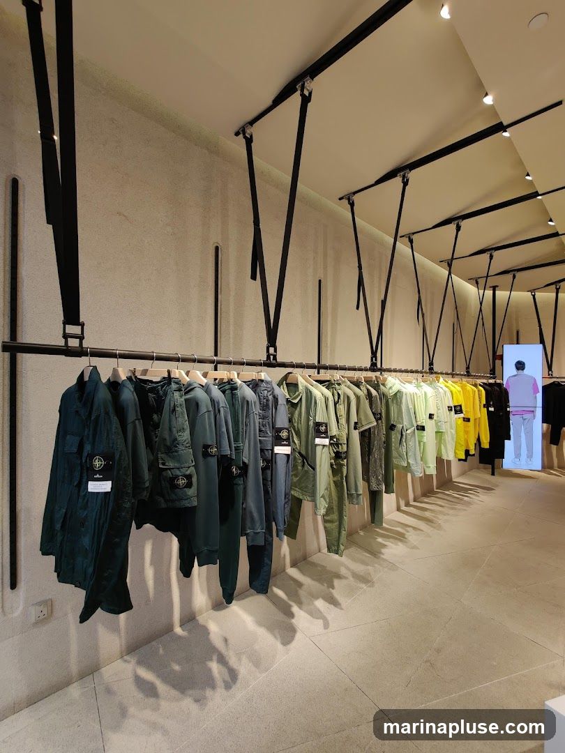 A row of men's clothing, primarily in shades of green, blue, and yellow, is displayed on hangers along a rack in a modern store. The garments include jackets, shirts, and pants, many featuring the Stone Island logo. The store has a minimalist design with textured walls and a suspended ceiling with track lighting. In the background, a digital display shows a mannequin wearing a similar outfit.