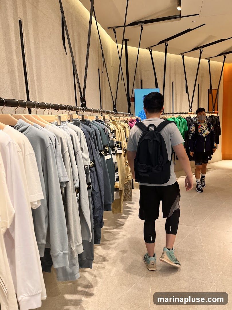 The image shows the interior of a clothing store with racks of apparel. Two men are visible, one walking towards the camera and another further down the aisle. The clothing appears to be casual wear, with a variety of colors and styles on display. The store has a modern aesthetic with minimalist decor and lighting.