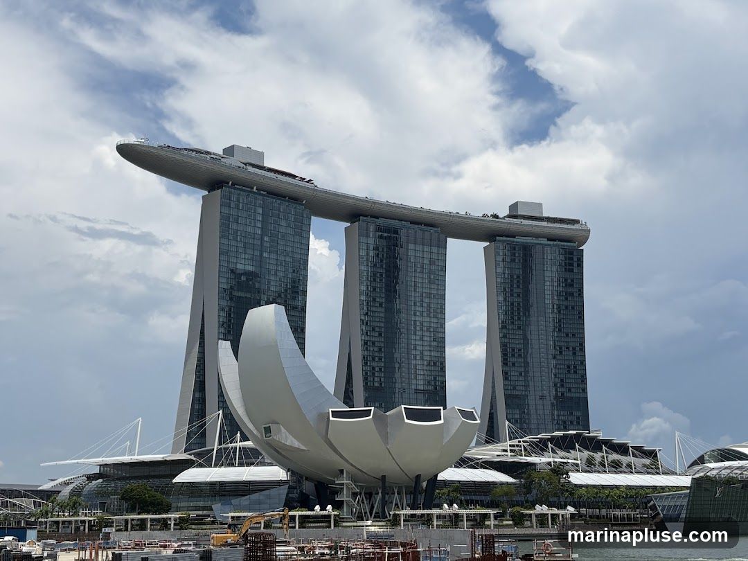 A wide shot of the Marina Bay Sands complex in Singapore under a cloudy sky. The iconic three towers, connected by the SkyPark, are visible, along with the ArtScience Museum in the foreground. Construction is ongoing in the lower part of the image.