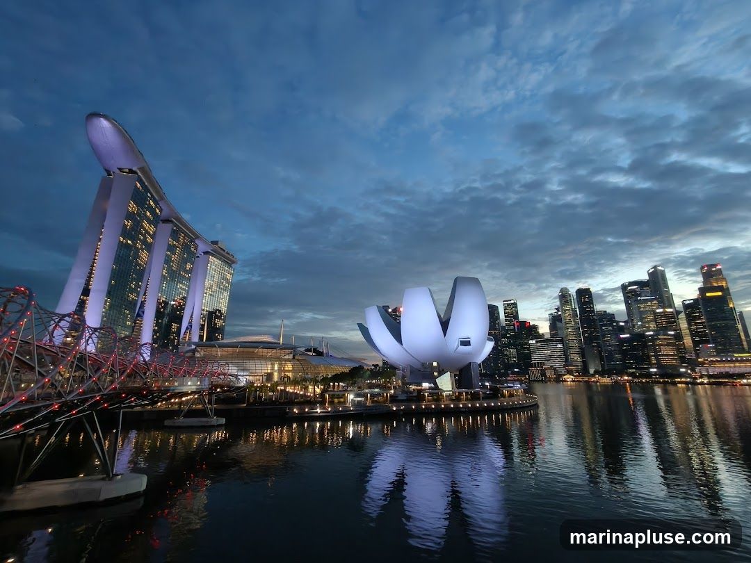 A wide-angle view of the Singapore skyline at dusk, featuring the Marina Bay Sands hotel, the ArtScience Museum (shaped like a lotus flower), and the Helix Bridge. The buildings are illuminated, and their reflections are visible in the calm water. The sky is a deep blue with dramatic clouds.
