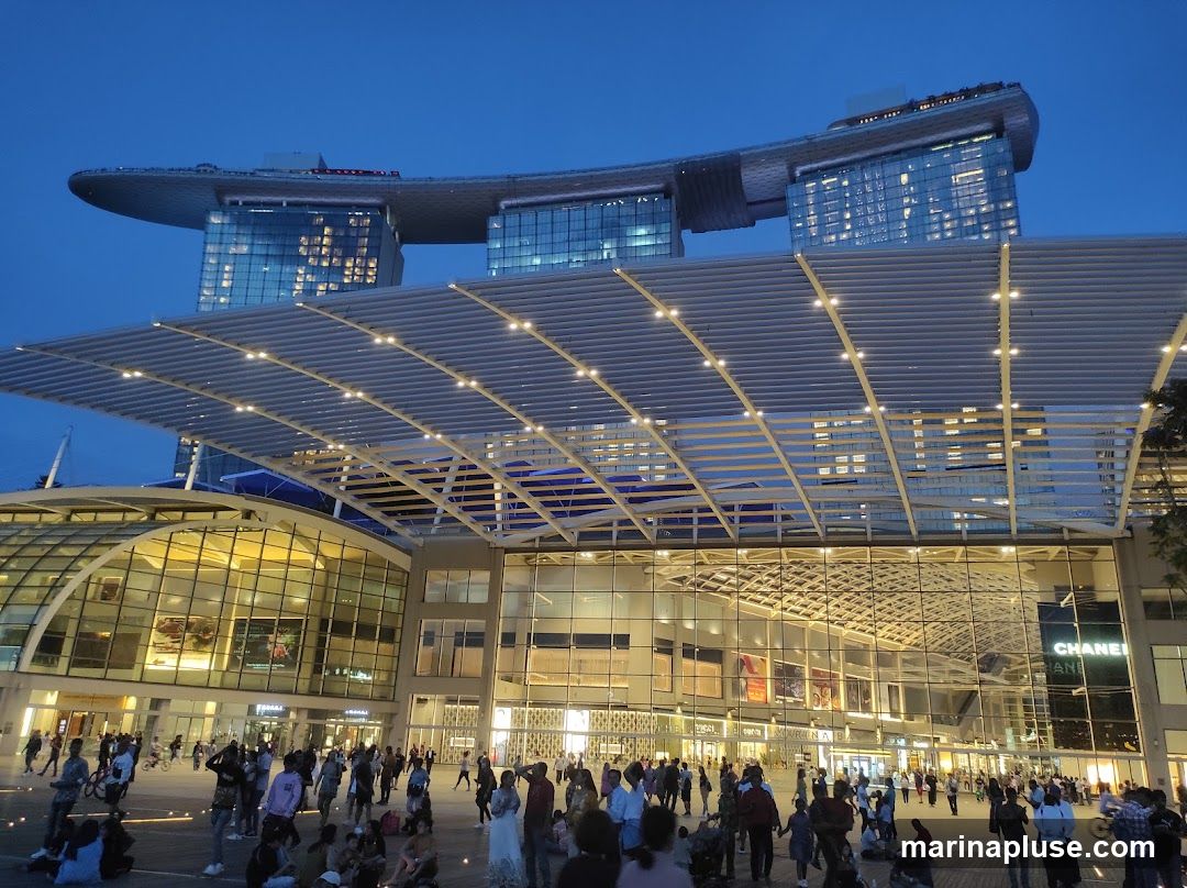 A wide shot of the Marina Bay Sands Shoppes complex in Singapore at dusk. The iconic three-tower hotel structure is visible in the background, illuminated against the twilight sky. In the foreground, a large, modern shopping mall with extensive glass facades is bustling with people. The mall features a distinctive, large, white, slatted canopy extending over the outdoor plaza. Signs for various shops, including CHANEL, are visible on the building's exterior. The plaza is filled with pedestrians, some sitting and others walking, enjoying the evening atmosphere.