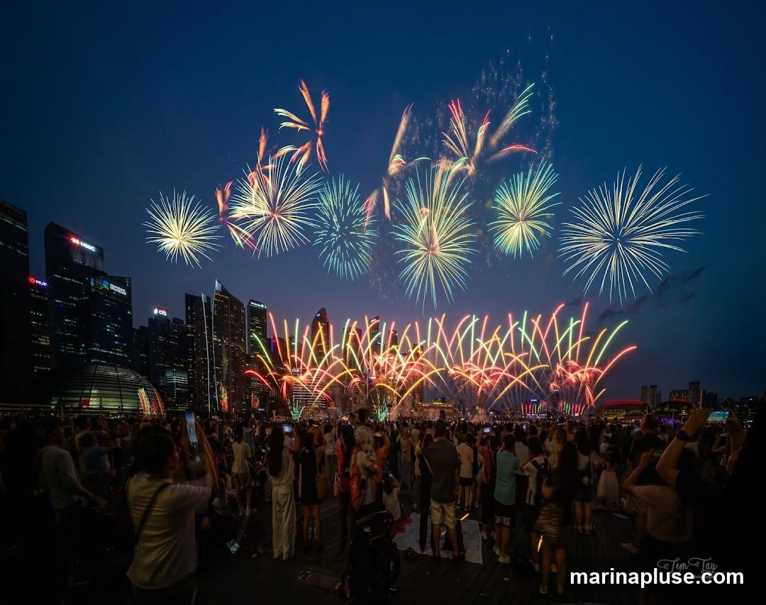 A vibrant fireworks display illuminates the night sky above a modern city skyline. A large crowd of people gathers on a waterfront promenade, many holding up their phones to capture the spectacle. The fireworks are a mix of bright bursts and cascading trails of color, set against a deep blue twilight sky. The city buildings in the background are silhouetted, with some lights visible. The overall scene captures a festive and celebratory atmosphere.