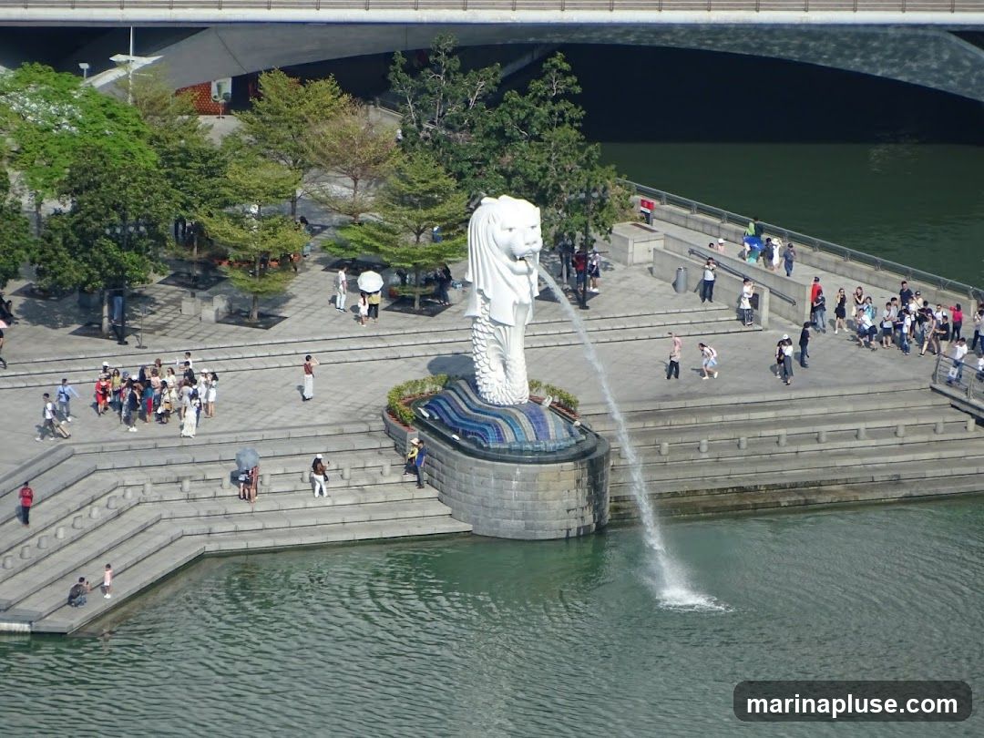 An aerial view of the Merlion fountain in Singapore, with tourists gathered around. The fountain is spraying water into the Singapore River.