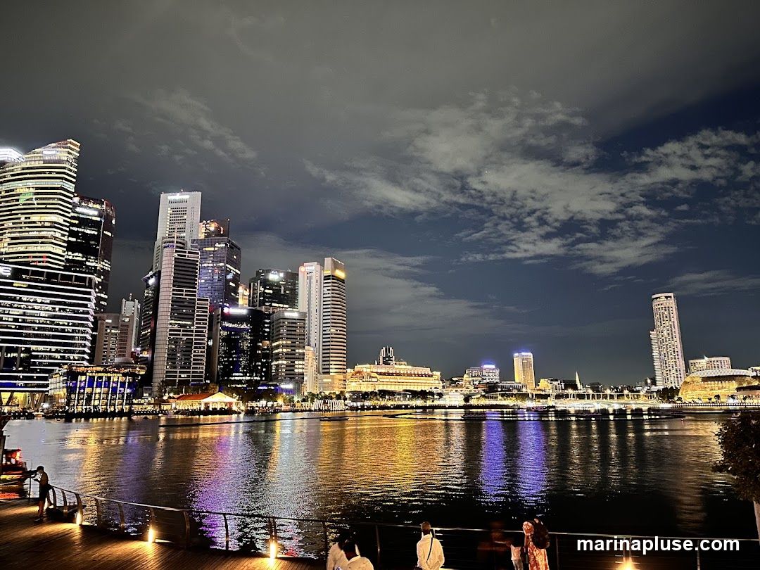 A wide shot of a city skyline at night, with numerous illuminated skyscrapers reflected in a body of water. The sky is dark with scattered clouds. In the foreground, a wooden boardwalk with a few people is visible.