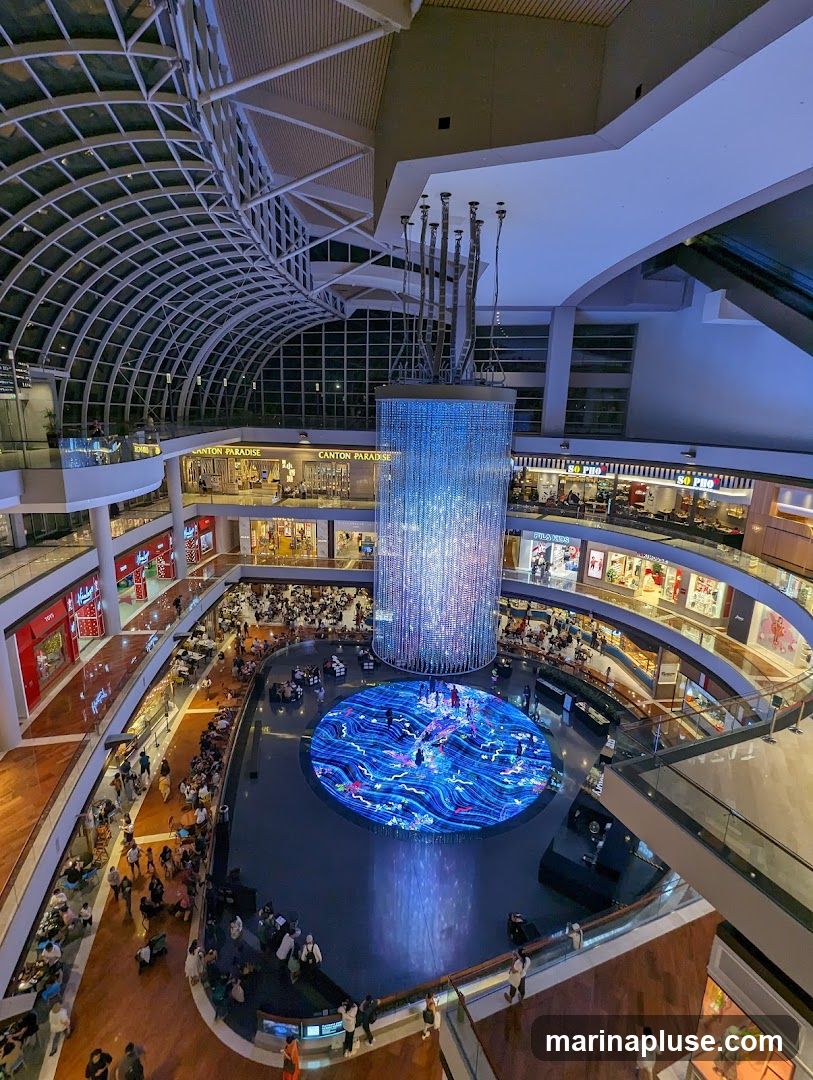 An aerial view of a multi-level indoor shopping mall. A large, circular water feature with a vibrant blue, animated display is at the center of the mall. Above it, a tall, cylindrical light installation sparkles with thousands of lights. Shoppers are seen walking on the various levels and seated around the water feature. Storefronts with visible signage are present on each level, including "CANTON PARADISE" and "SO PHO". The mall has a modern architectural design with a curved glass ceiling.