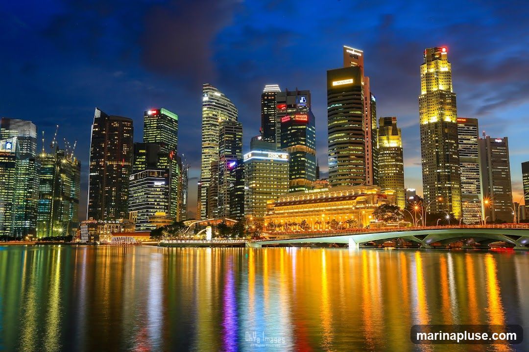 A panoramic view of the Singapore skyline at night, with illuminated skyscrapers reflecting in the calm waters of a bay. The sky is a deep blue with hints of clouds, and the city lights create a vibrant spectacle. A bridge spans across the water in the foreground, connecting different parts of the city. The architecture is modern and diverse, showcasing a bustling urban landscape.