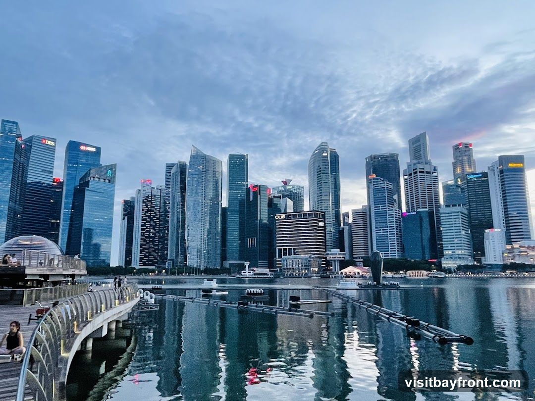 A panoramic view of the Singapore skyline at dusk, with modern skyscrapers reflecting in the calm waters of Marina Bay. A curved walkway with people strolling and sitting is in the foreground, leading towards a domed structure. The sky is filled with dramatic clouds, hinting at the transition from day to night.