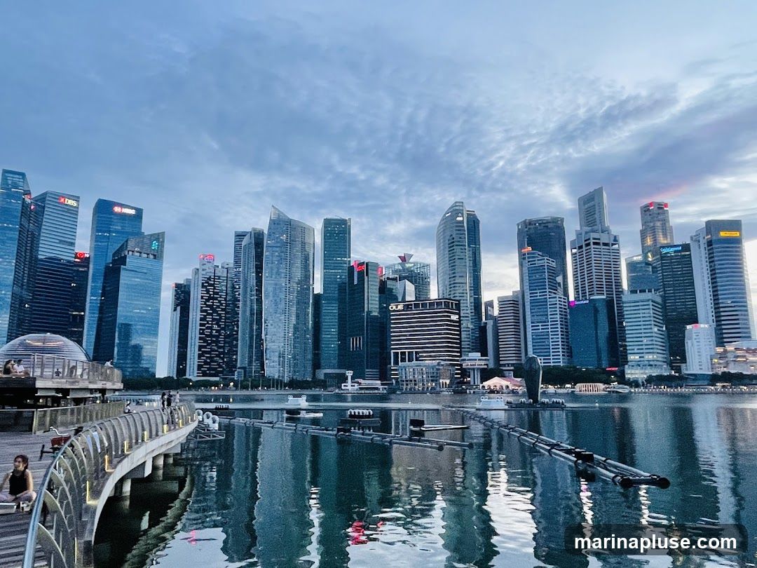 A panoramic view of the Singapore skyline at dusk, with modern skyscrapers reflecting in the calm waters of Marina Bay. A curved walkway with people strolling and sitting is in the foreground, leading towards a domed structure. The sky is filled with dramatic clouds, hinting at the transition from day to night.