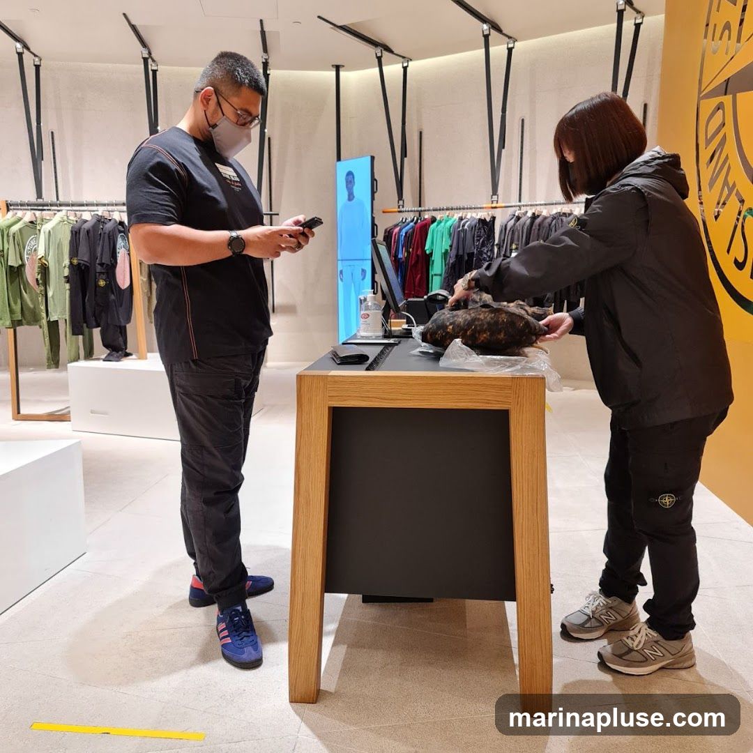 A man wearing a mask and a black t-shirt stands holding a phone, looking at it. A woman in a black jacket is at a wooden counter, holding a dark, textured item. Racks of clothing are visible in the background, along with a large yellow circular logo on the right side of the image. The store appears to be a retail clothing store.