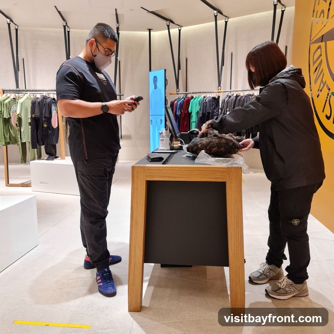 A man wearing a mask and a black t-shirt stands holding a phone, looking at it. A woman in a black jacket is at a wooden counter, holding a dark, textured item. Racks of clothing are visible in the background, along with a large yellow circular logo on the right side of the image. The store appears to be a retail clothing store.
