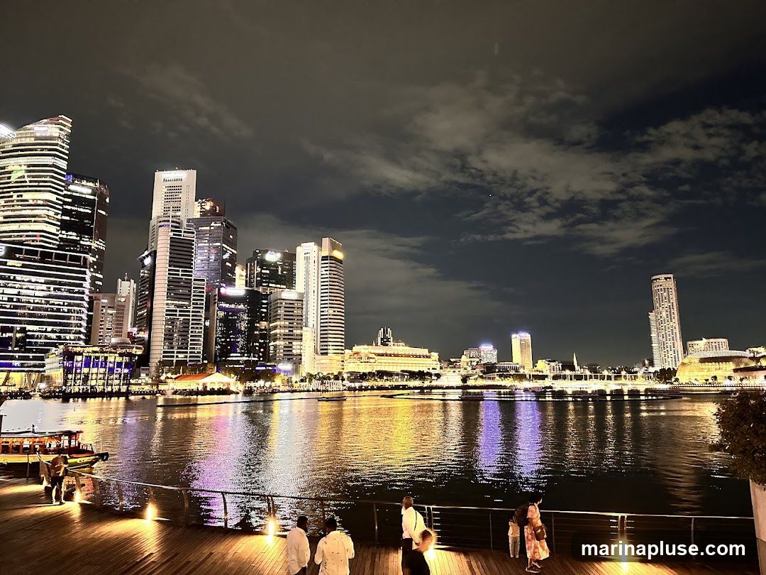 A wide shot of a city skyline at night, with numerous illuminated skyscrapers reflecting in the water. Several people are visible on a wooden boardwalk in the foreground, looking out at the view. Boats are also present on the water. The sky is dark with scattered clouds.