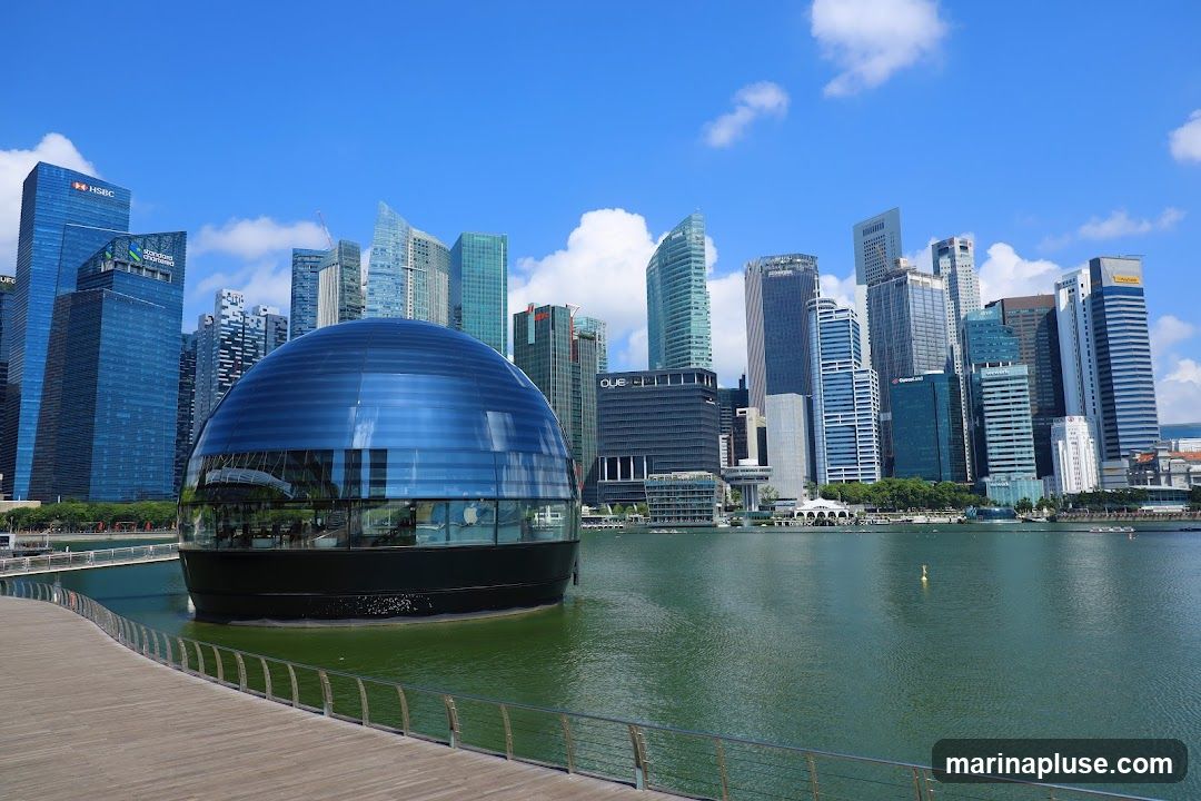 A wide shot of a futuristic, spherical building with a blue glass facade, situated in a body of water. The building is surrounded by a modern cityscape of tall skyscrapers under a bright blue sky with scattered clouds. A wooden walkway with a metal railing curves along the foreground, leading towards the water and the unique structure.
