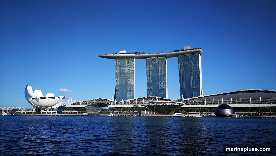 A wide shot of the Marina Bay Sands complex in Singapore, featuring the iconic three towers connected by a rooftop deck, and the ArtScience Museum with its lotus-like design. The buildings are set against a clear blue sky, with the dark blue water of the bay in the foreground.