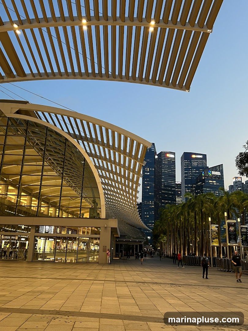 A wide-angle view of a modern building with a distinctive curved, slatted roof structure, set against a backdrop of skyscrapers. The building's facade features large glass windows, revealing illuminated interiors. A paved walkway leads away from the building, lined with palm trees and several people walking. The sky is clear and blue, indicating either early morning or late afternoon. Signs are visible on poles along the walkway, and building names like HSBC and DBS are discernible on the distant skyscrapers.