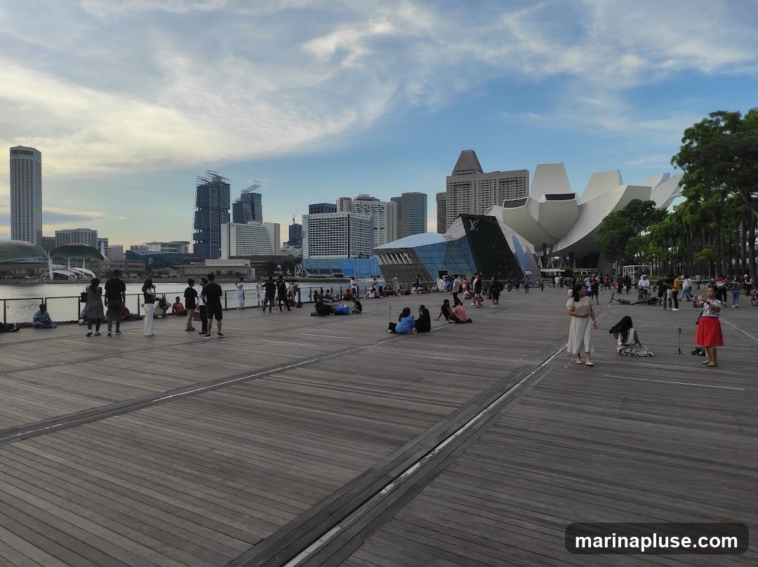 A wide wooden boardwalk is filled with people sitting and standing, enjoying the view of a city skyline. The skyline features modern skyscrapers and a distinctive lotus-shaped building. The sky is partly cloudy with a mix of blue and white.