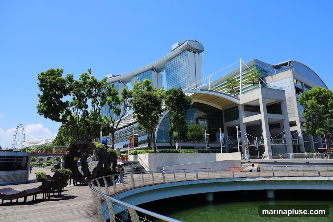A wide shot captures the impressive modern architecture of a large building complex, possibly a hotel or resort, with its distinctive sky-bridge connecting two towers. The building features extensive glass facades and a curved, canopy-like roof structure over a lower section. In the foreground, a wooden boardwalk curves over a green body of water, with a metal railing. Lush green trees are scattered throughout the scene, and a Ferris wheel is visible in the distance. The sky is clear and blue.