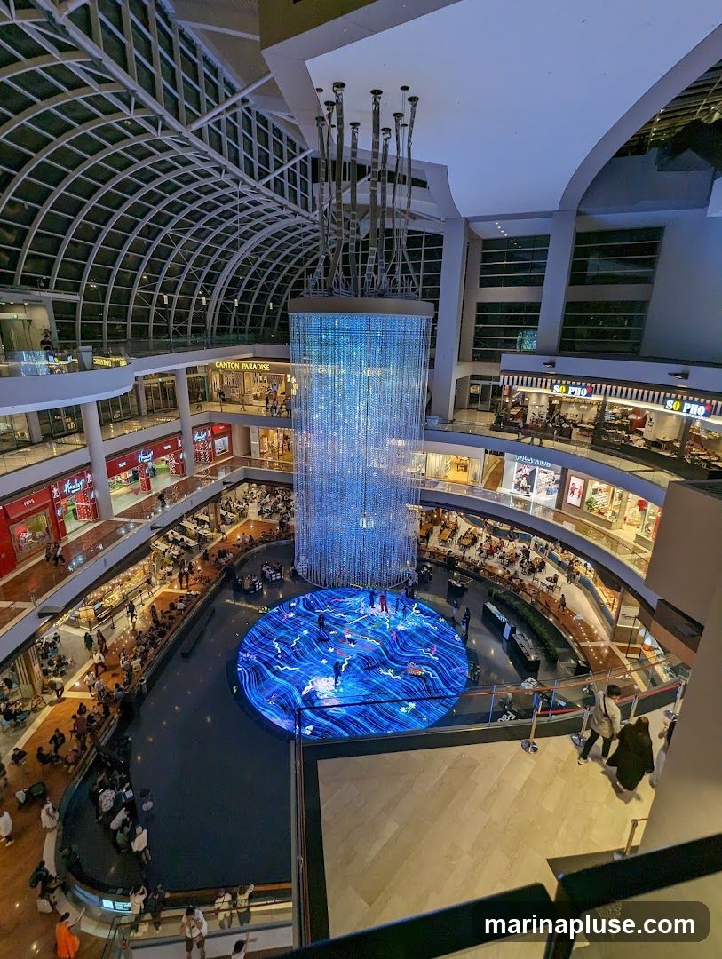 The image shows the interior of a multi-level shopping mall. A large, circular water feature is in the center, with a digital display on its surface showing a blue, wave-like pattern with figures walking on it. Above the water feature is a tall, cylindrical light installation made of numerous hanging strands of lights, creating a shimmering effect. People are seen walking around the mall, browsing shops, and sitting at tables. The mall has a modern design with a glass ceiling and multiple balconies overlooking the central area. Shops with visible signage like 'Canton Paradise' and 'So Pho' are present on different levels.
