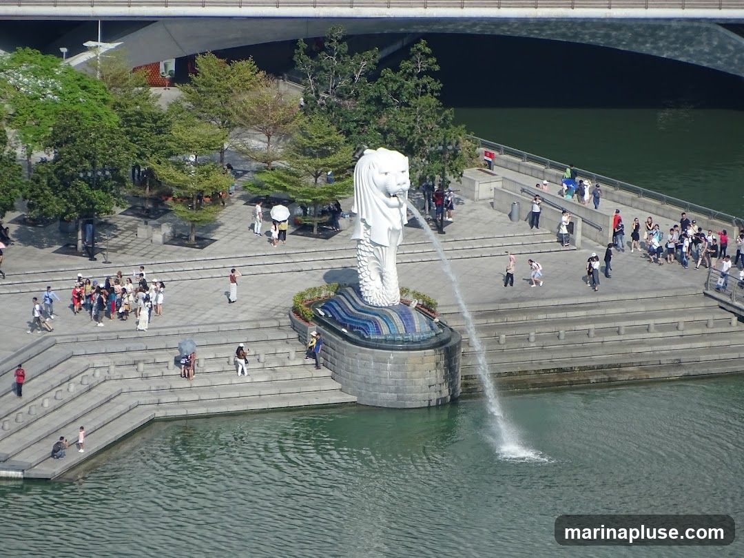 An aerial view of the Merlion statue in Singapore, with water spouting from its mouth into the Singapore River. Many tourists are gathered around the statue and along the waterfront promenade. Trees and a modern bridge are visible in the background.