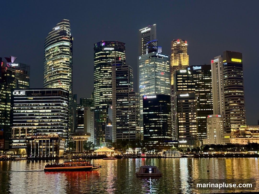 A nighttime view of the Singapore skyline, featuring numerous illuminated skyscrapers reflected in the water. A boat is visible on the water in the foreground.