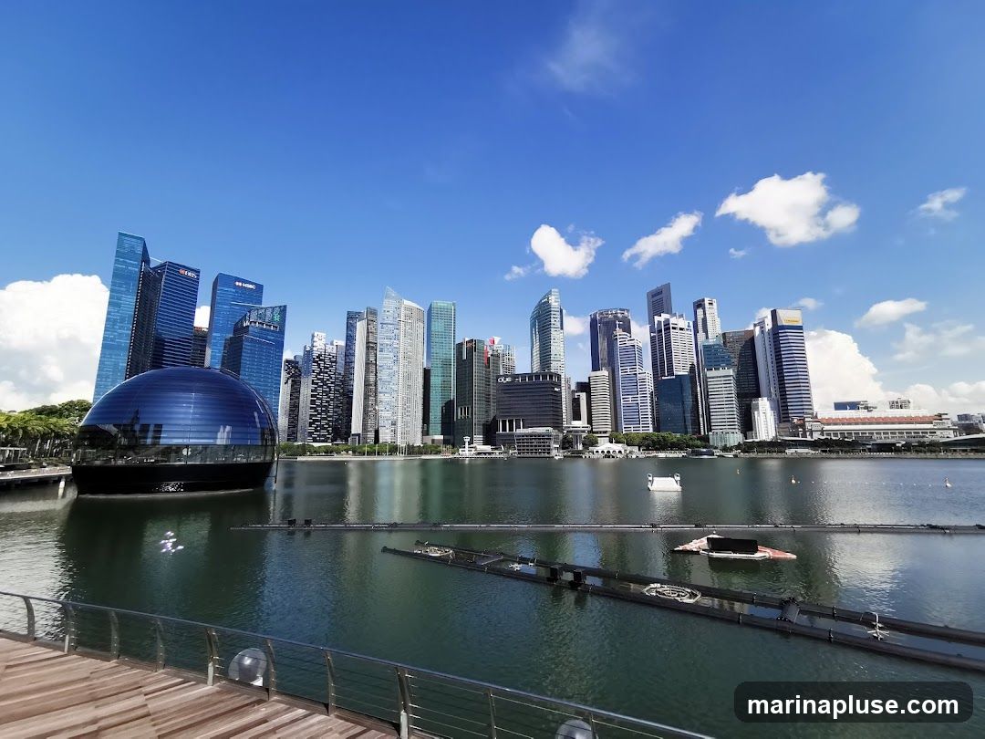A panoramic view of the Singapore skyline, featuring numerous modern skyscrapers and the distinctive Marina Bay Sands resort. The image captures the bustling cityscape reflected in the calm waters of Marina Bay under a bright blue sky with scattered clouds. In the foreground, a wooden walkway with a railing overlooks the bay.