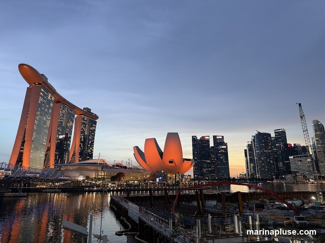 A wide shot of the Singapore skyline at dusk, featuring the Marina Bay Sands hotel and the ArtScience Museum. The buildings are illuminated, casting reflections on the water. In the foreground, there is a construction site with cranes and scaffolding. The sky is a gradient of blue and orange.