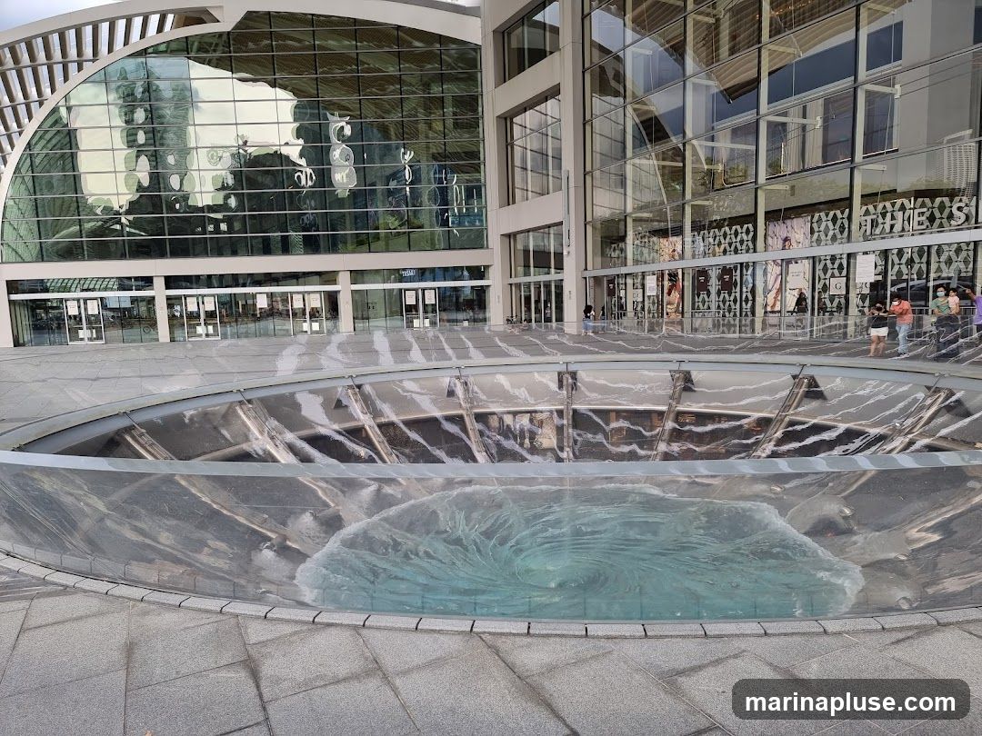 A wide shot of a modern building with a large glass facade and a circular water feature in the foreground. The water feature has a swirling vortex of blue water. The building's reflection is visible in the glass. Several people are standing near the entrance of the building.