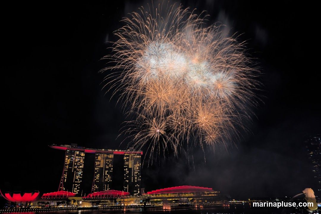 A vibrant fireworks display illuminates the night sky over Singapore's Marina Bay Sands hotel and the surrounding cityscape. The iconic three-towered hotel, illuminated with red and gold lights, stands prominently on the left, with its distinctive boat-shaped rooftop visible. Below, the ArtScience Museum, shaped like a lotus flower, also glows red. The Merlion statue, a mythical creature with a lion's head and a fish's body, is visible on the right, spouting water. The fireworks are a mix of golden, white, and orange bursts, creating a dazzling spectacle against the dark sky.