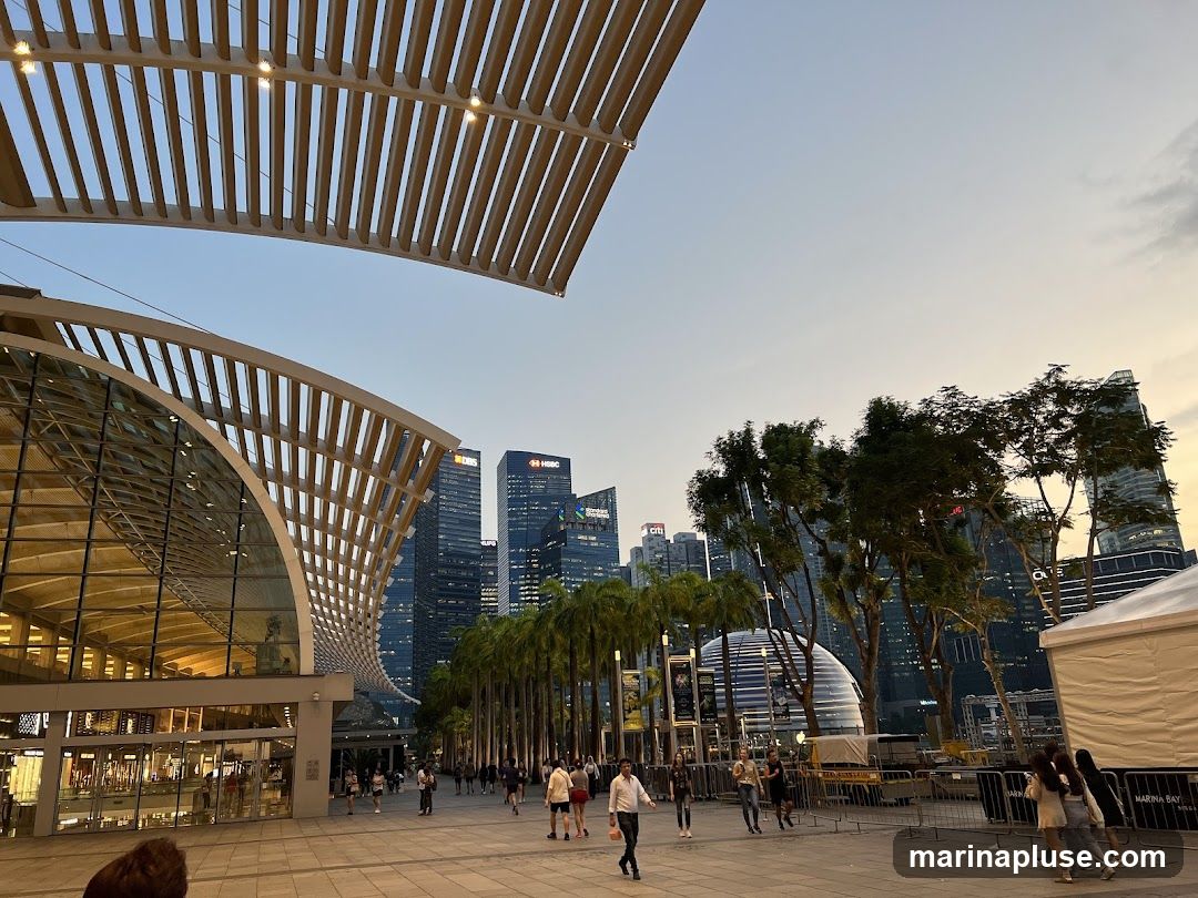 A wide shot of a modern architectural complex with a curved, slatted roof extending over a glass-fronted building. In the background, a cityscape of tall skyscrapers, including buildings with HSBC and DBS logos, is visible under a pale sky. Palm trees line a walkway where people are strolling. A white tent structure is on the right side of the image.