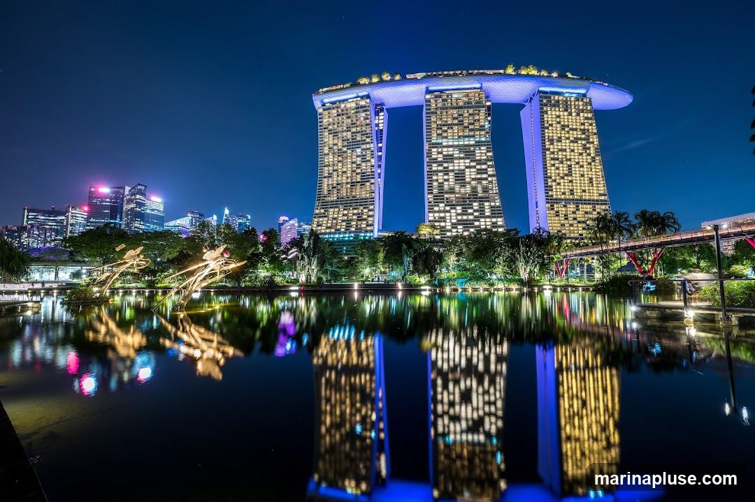 A nighttime view of the Marina Bay Sands hotel in Singapore, with its iconic three towers and sky park illuminated. The building's lights are reflected in the calm water of a nearby bay. The cityscape of Singapore is visible in the background, along with lush greenery and decorative lights in the foreground.