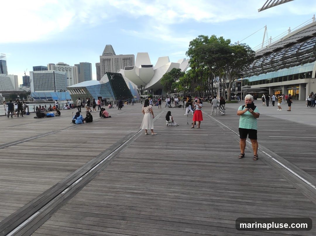 A wide wooden boardwalk is filled with people relaxing and walking. In the background, a modern city skyline features distinctive architecture, including a building with a lotus-like design and a glass-fronted structure. The sky is partly cloudy.