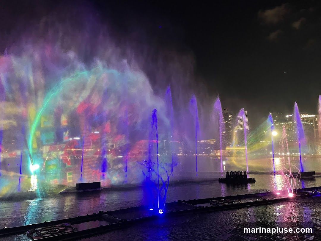 A vibrant night-time water and light show with colorful fountains and projections on mist, set against a city skyline.