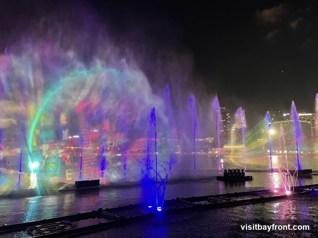A vibrant night-time water and light show with colorful fountains and projections on mist, set against a city skyline.