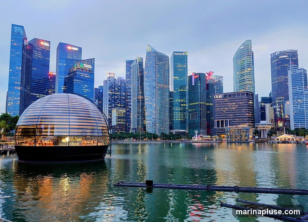 A panoramic view of the Singapore skyline at dusk, featuring the iconic Marina Bay Sands hotel and a cluster of modern skyscrapers. In the foreground, a unique spherical glass building, likely a retail establishment, sits on the water. The buildings are illuminated, reflecting on the calm bay.