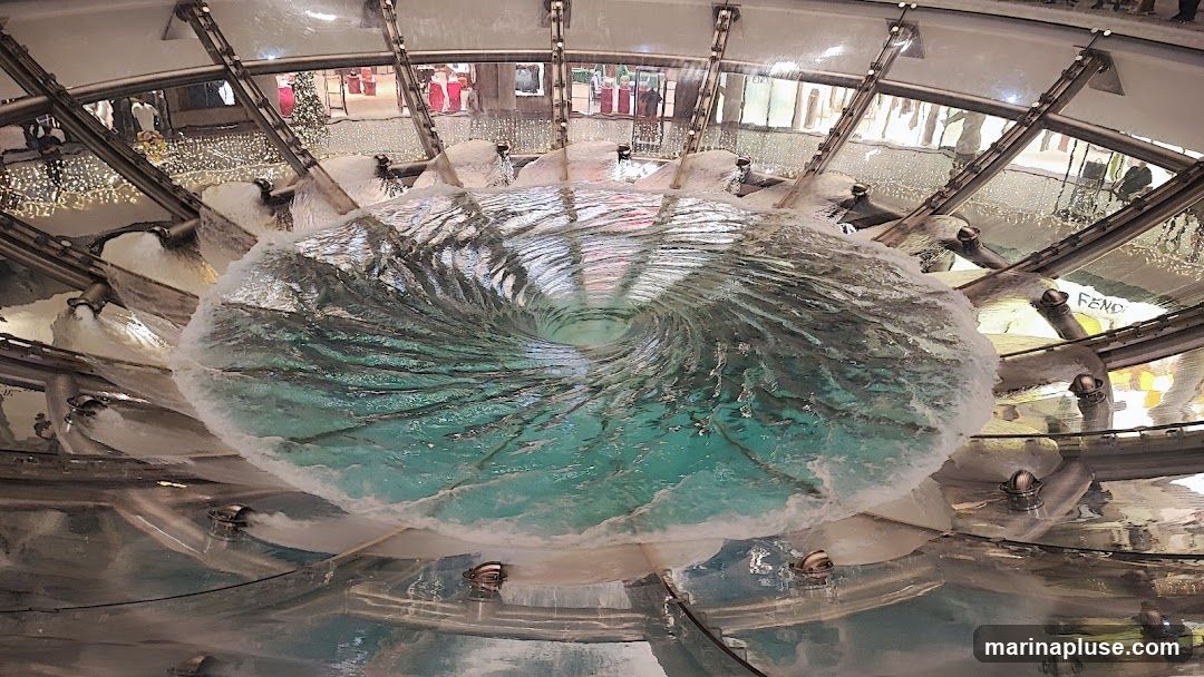 A close-up, fisheye view of a large, circular fountain with water swirling into a vortex in the center. The water is a vibrant turquoise color and appears to be illuminated from below. The fountain is surrounded by a metallic structure with pipes and nozzles, and in the background, reflections of shops and holiday decorations can be seen. One reflection clearly shows the Fendi logo.