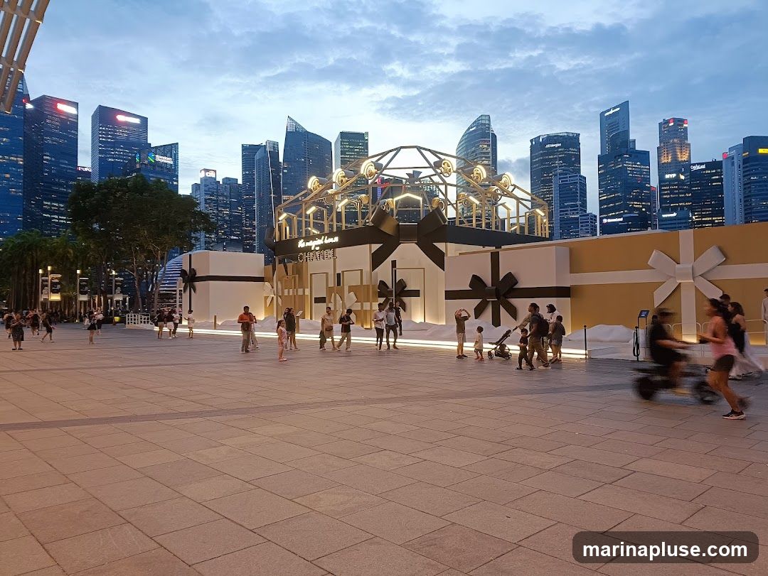 A large outdoor display resembling gift boxes, with the Chanel logo prominently featured. People are walking around the display in a plaza with a city skyline in the background. The sky is overcast and appears to be dusk.