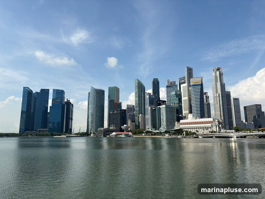 A panoramic view of a modern city skyline with numerous skyscrapers of varying heights and architectural styles, reflected in the calm water of a bay or river in the foreground. The sky is blue with scattered white clouds. Several buildings have visible corporate logos such as HSBC, UOB, and FWD.