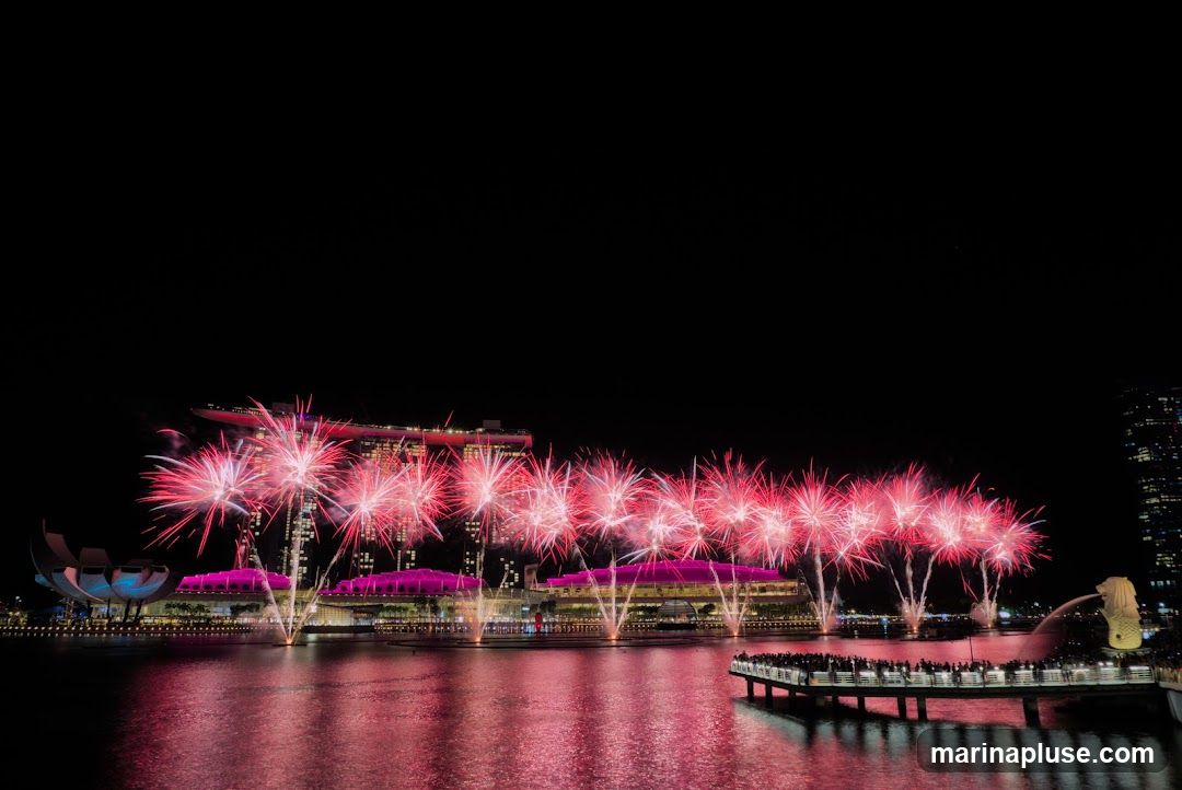 A vibrant fireworks display illuminates the night sky over Marina Bay in Singapore. The fireworks, predominantly in shades of pink and white, erupt in a dazzling spectacle above the iconic Marina Bay Sands hotel and the ArtScience Museum. The reflection of the fireworks and the illuminated buildings shimmers on the calm waters of the bay. A pier filled with spectators can be seen in the foreground, observing the event.