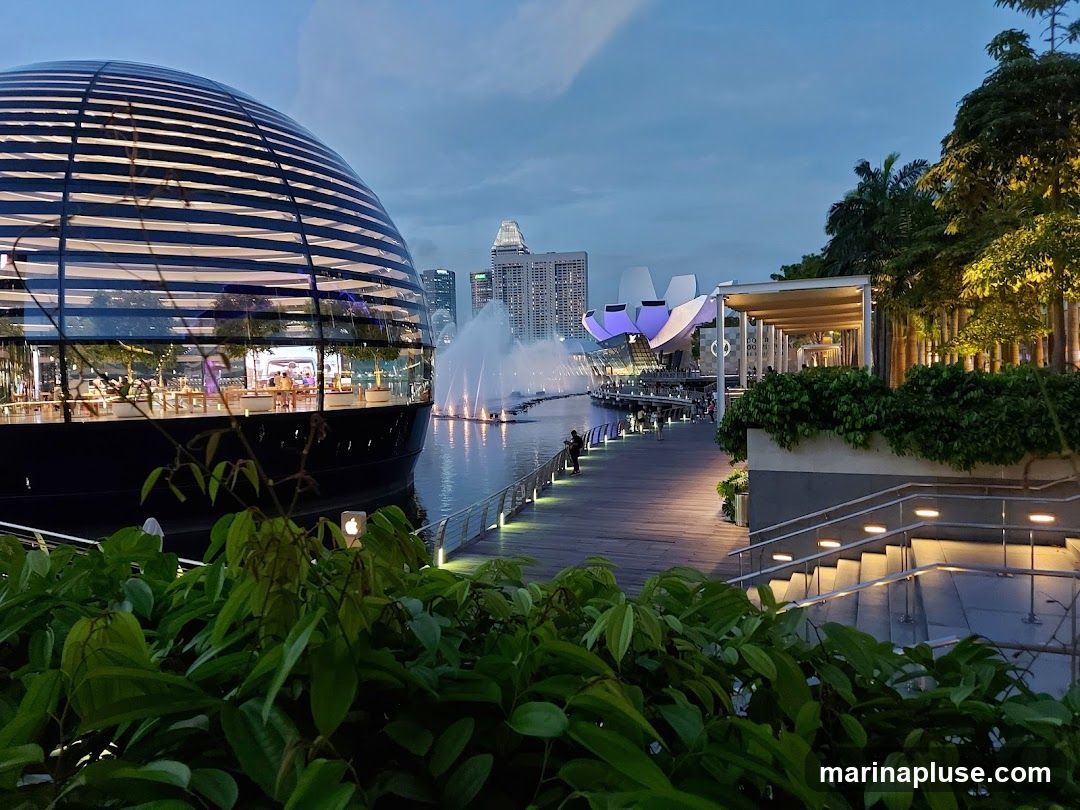 A wide shot of a waterfront promenade at dusk, featuring a distinctive spherical glass building with internal lighting and trees. In the background, a fountain sprays water, and modern city buildings rise against the twilight sky. To the right, a covered walkway with lush greenery and illuminated stairs leads away from the water. The scene conveys a sense of urban leisure and contemporary design.