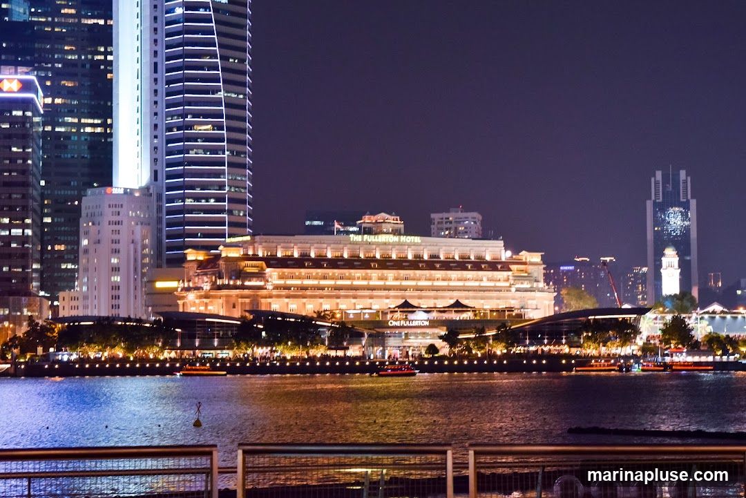 A nighttime view of the Fullerton Hotel in Singapore, illuminated by warm lights. The hotel is a grand, classical building situated along the waterfront, with modern skyscrapers visible in the background. Boats are on the water in front of the hotel.