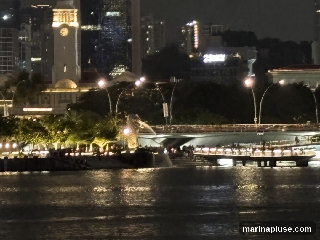 A nighttime cityscape featuring a prominent clock tower, modern skyscrapers, and a famous statue of a mythical creature spitting water. A bridge spans across the water in the foreground, with people visible on a promenade below. The water reflects the city lights.