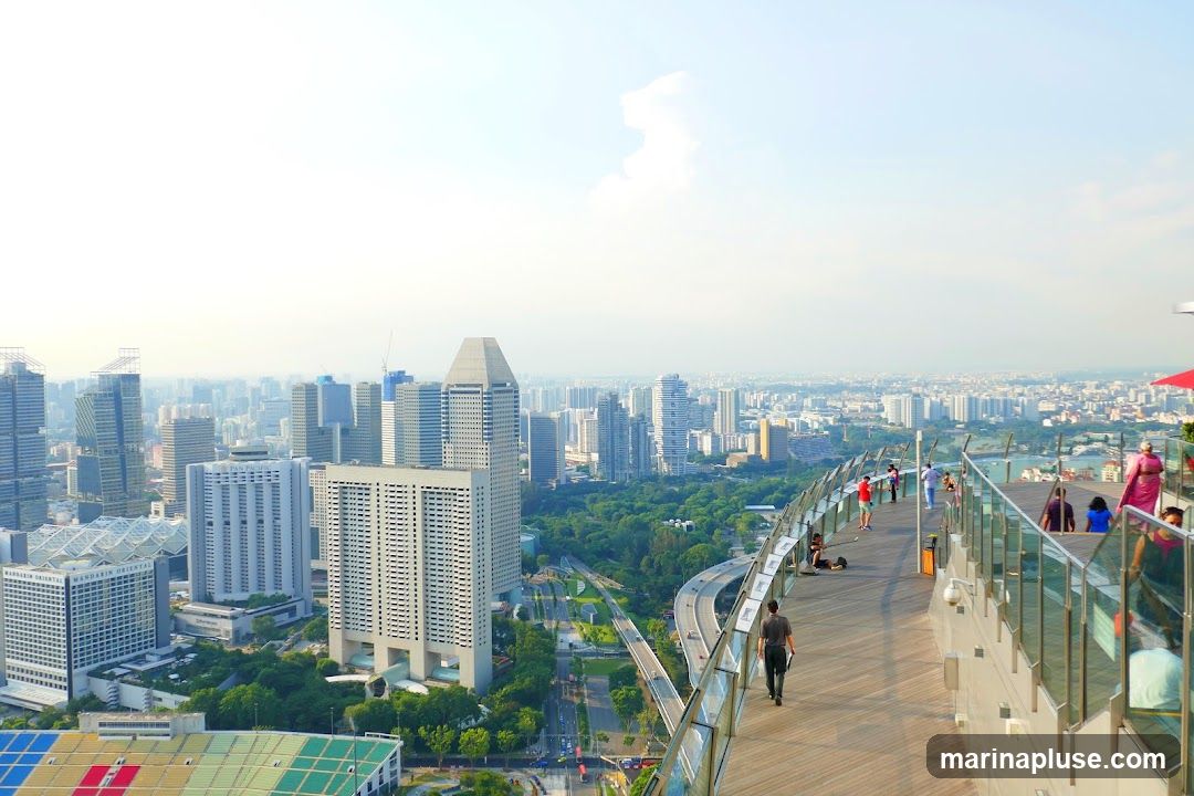 A panoramic view of a dense cityscape with numerous skyscrapers and buildings, seen from a high vantage point. In the foreground, a wooden observation deck with glass railings curves along the edge, with several people strolling and relaxing. Lush green trees are visible between the buildings, and a highway system winds through the urban landscape. The sky is bright and hazy.