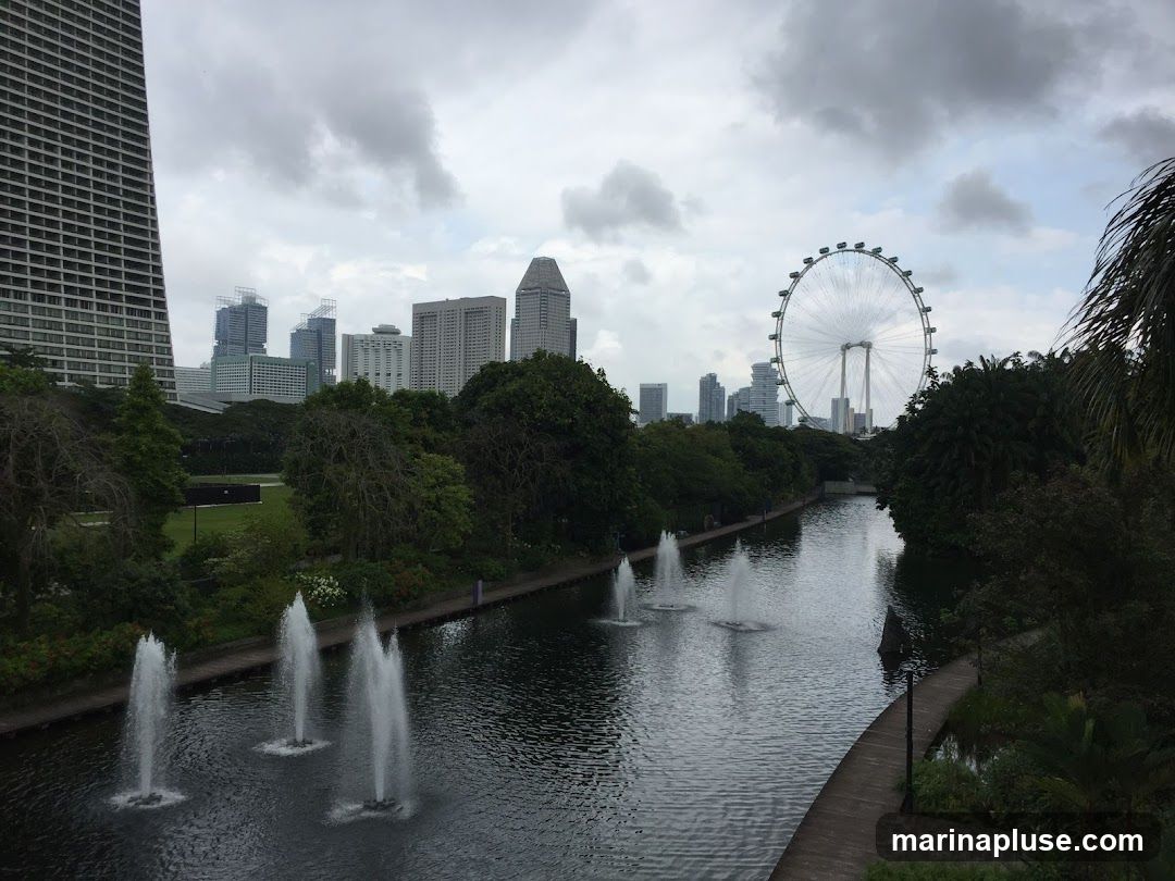 A wide shot of a city skyline under a cloudy sky, featuring a tall skyscraper on the left, a river with fountains in the foreground, and a large Ferris wheel in the background. Lush green trees and foliage line the riverbanks, with a wooden walkway visible on the right. The water reflects the overcast sky and the surrounding greenery.