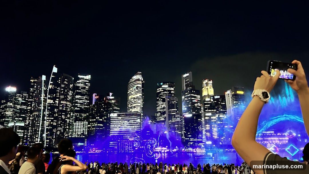 A crowd of people watches a vibrant light and water show against a backdrop of illuminated skyscrapers at night. One person in the foreground is taking a photo of the spectacle with their smartphone. The buildings are lit with countless windows, and the light show features blue and purple hues, with water jets and geometric light patterns.