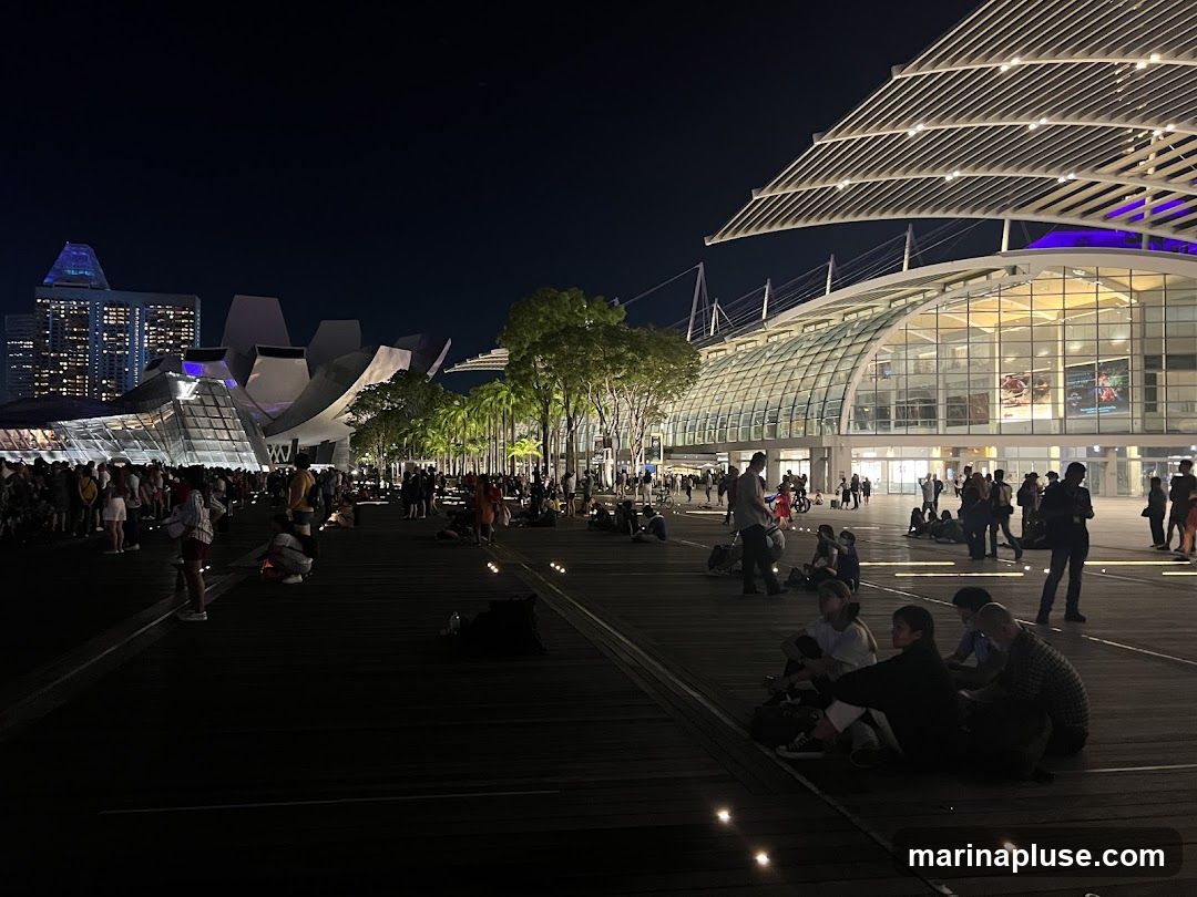A crowd of people are gathered on a wooden boardwalk at night, in front of a large, modern building with a glass facade and a distinctive white, ribbed roof structure. In the background, a cityscape with illuminated skyscrapers is visible. The atmosphere appears relaxed, with many people sitting on the ground, some in groups, while others stand and walk around. The lighting is a mix of ambient city lights, spotlights on the building, and small lights embedded in the boardwalk.