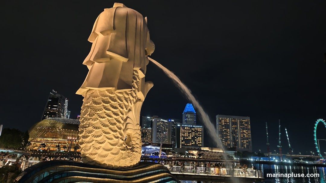 A night view of the Merlion statue in Singapore, with water spouting from its mouth. In the background, the city skyline is illuminated, featuring modern skyscrapers, the Esplanade, and the Singapore Flyer.