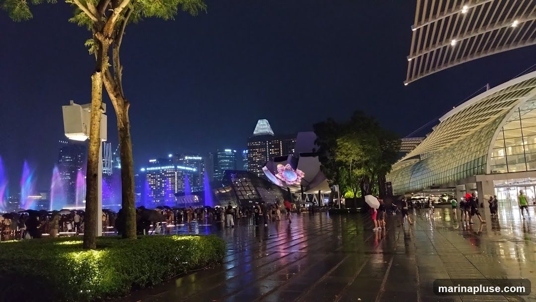 A nighttime view of a city skyline featuring illuminated buildings and a water fountain show. In the foreground, a wet promenade reflects the colorful lights. People are seen walking with umbrellas, suggesting it might be raining. Modern architectural structures, including a building with a distinctive curved roof, are visible on the right. The overall scene is vibrant and dynamic, with a blend of urban landscape and entertainment.