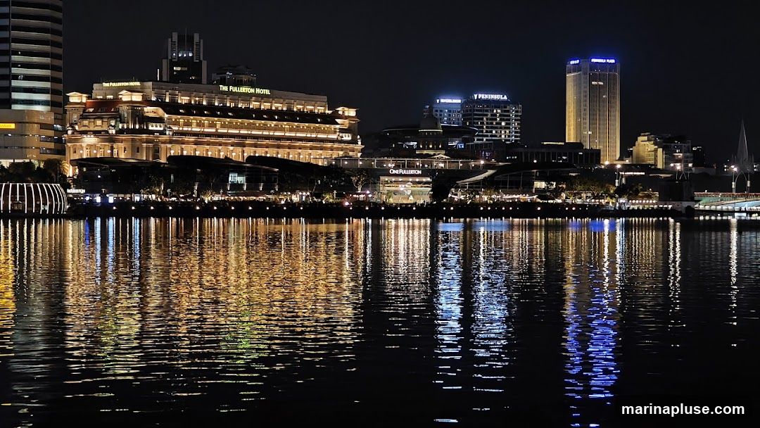 A nighttime view of a city skyline across a body of water. The buildings are illuminated, casting reflections on the dark water. Prominent buildings include The Fullerton Hotel and Peninsula Plaza. The scene is characterized by the contrast between the dark sky and the bright lights of the city and their reflections.