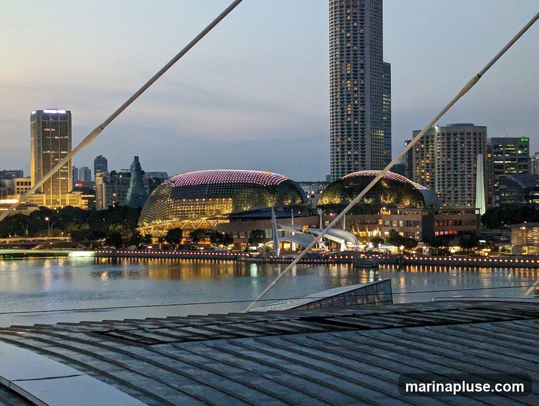 A panoramic view of a city skyline at dusk, with a body of water in the foreground. Several modern skyscrapers and distinctive domed buildings are illuminated, reflecting on the water. Cables from a structure in the foreground stretch across the scene.