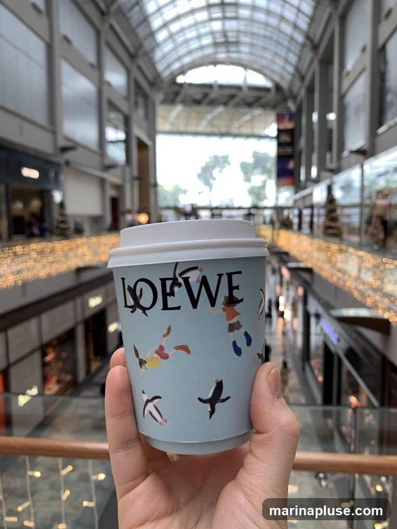 A hand holds a light blue disposable coffee cup with the Loewe logo and a whimsical illustration of penguins and people. The background shows a blurred interior of a modern shopping mall with arched ceilings and festive lights.