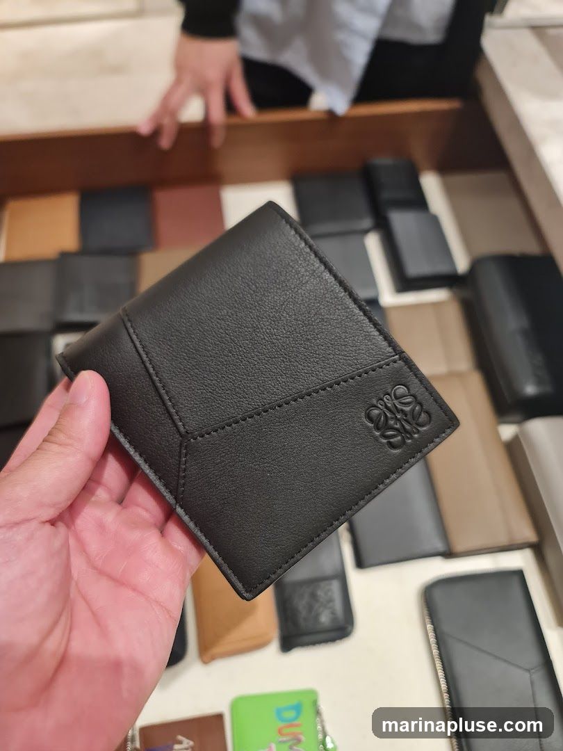 A hand holds a black leather wallet with a debossed logo. The wallet is displayed in a drawer filled with other leather goods, including wallets and cardholders, in various colors and styles.